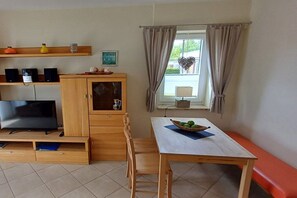 Een knus woongedeelte met een televisie, houten meubels, een eettafel met een schaal fruit en een raam met gordijnen.