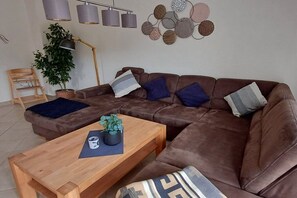 Een bruine leren hoekbank met blauwe en grijze kussens, een houten salontafel met een plant en een wandlamp.