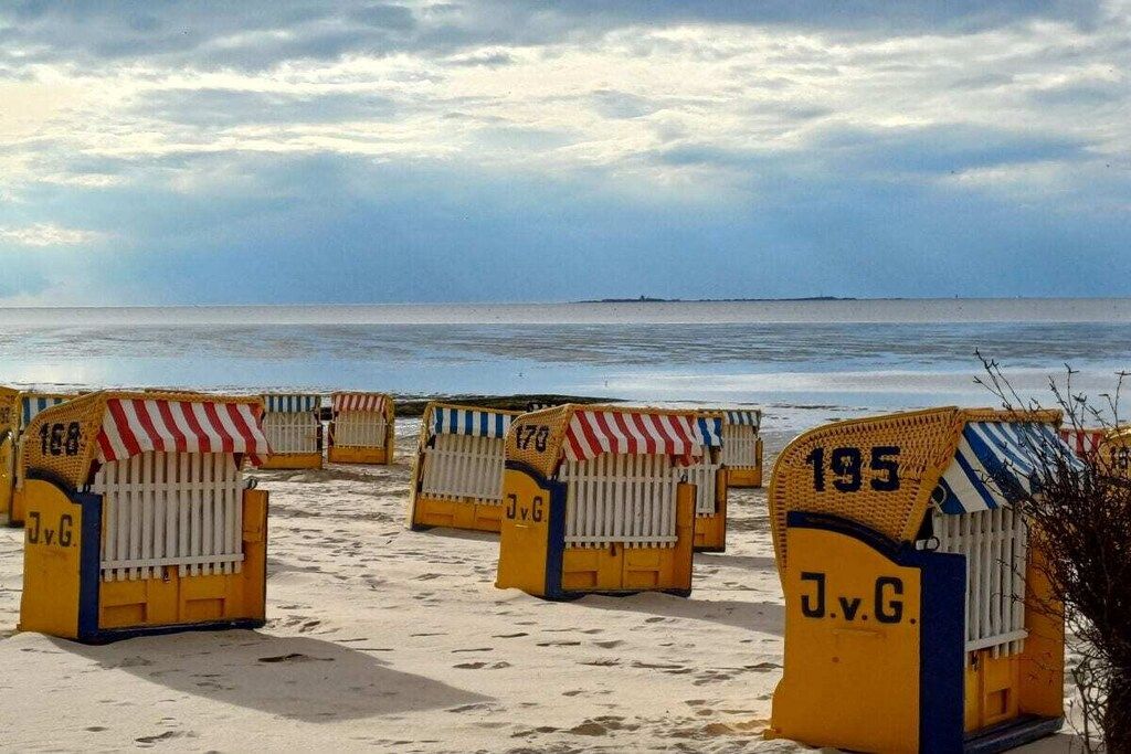 Des cabanes de plage portant des numéros et les initiales « J.v.G. », disposées sur une plage de sable, avec l’océan en arrière-plan.