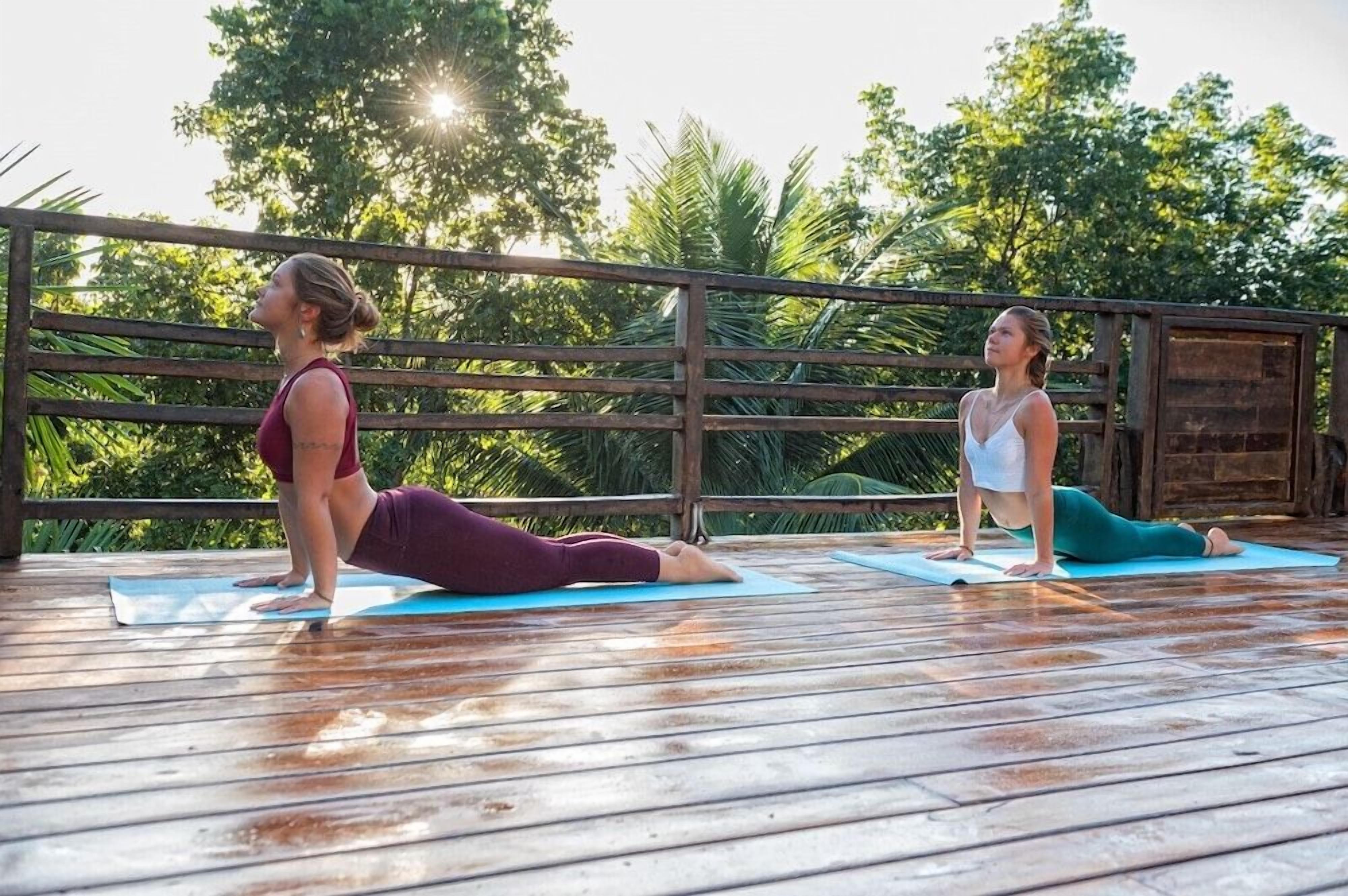 Two individuals on yoga mats on a wooden deck with a wooden railing and trees in the background.