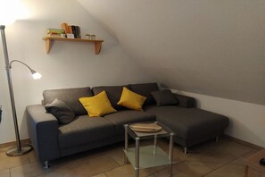 A modern living room with a sectional sofa, a glass coffee table, a floor lamp, and a wooden shelf with books and plants. - House on Häfchenweg Apartment 6 (Cuxhaven)