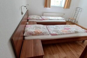 Una habitació amb dos llits de fusta, cadascun amb un coixí i una manta. Hi ha un finestral amb cortines, un radiador i una escala metàl·lica adossada a la paret.