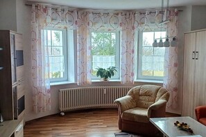 Una habitació amb sòl de fusta, un butac armchair beix, una taula amb un ram i una finestra amb cortines.