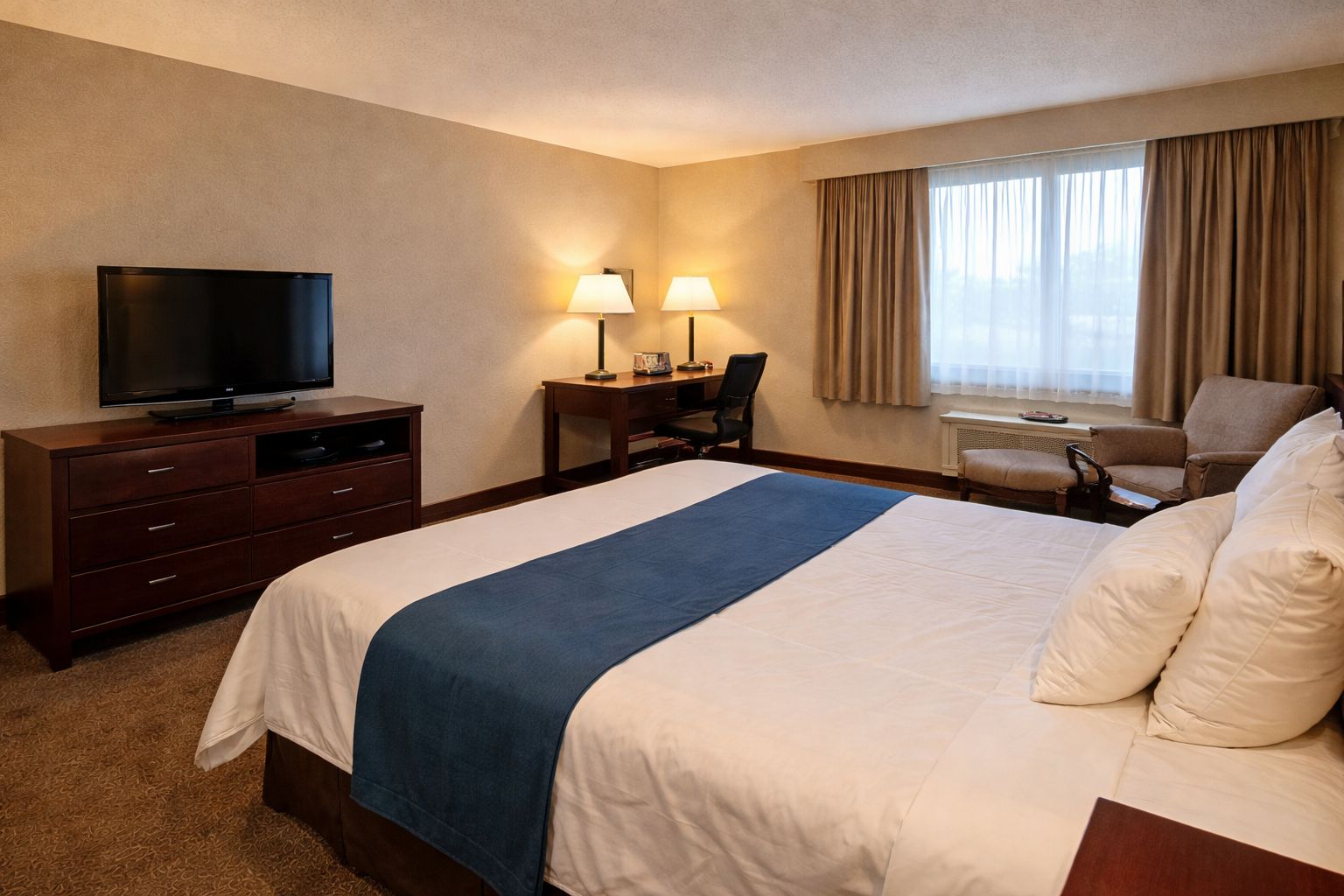 Una habitación de hotel con cama, escritorio con silla, televisor y ventana con cortinas.