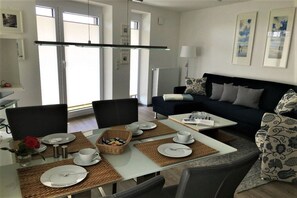 Una sala de estar moderna con sofá, mesa de centro, juego de comedor con platos y tazas, y vistas al exterior a través de amplias ventanas.