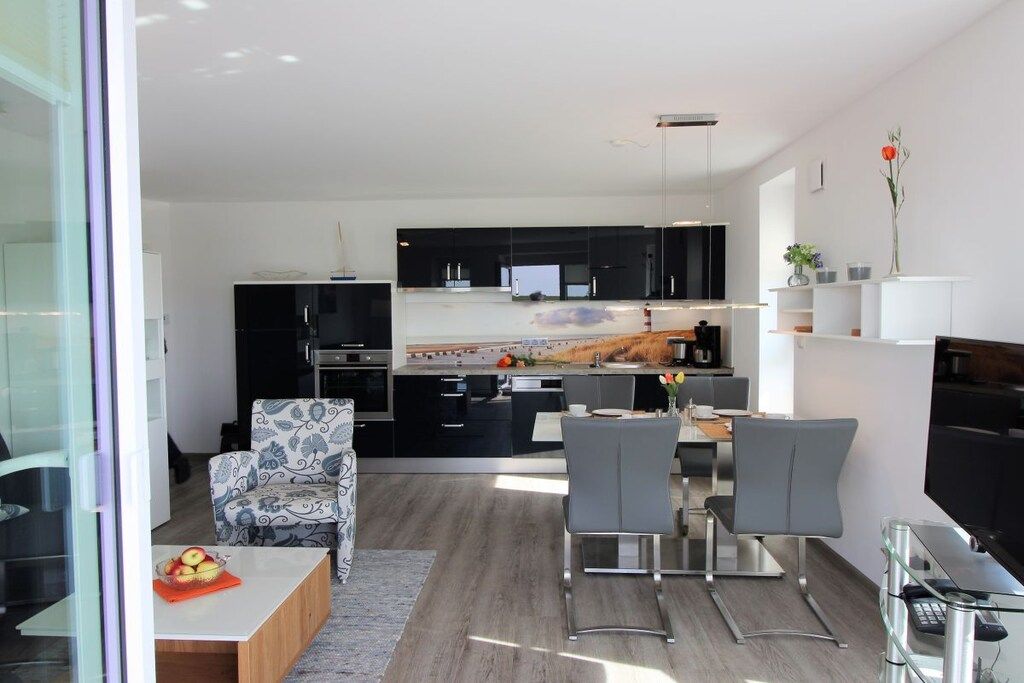 Una cocina moderna con armarios negros, un comedor con mesa de vidrio y un espacio de estar con un sillón estampado.