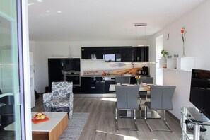A modern kitchen with black cabinets, a dining area with a glass table, and a living space with a patterned armchair. - Holiday apartment with 2 bedrooms (Dorum-Neufeld)