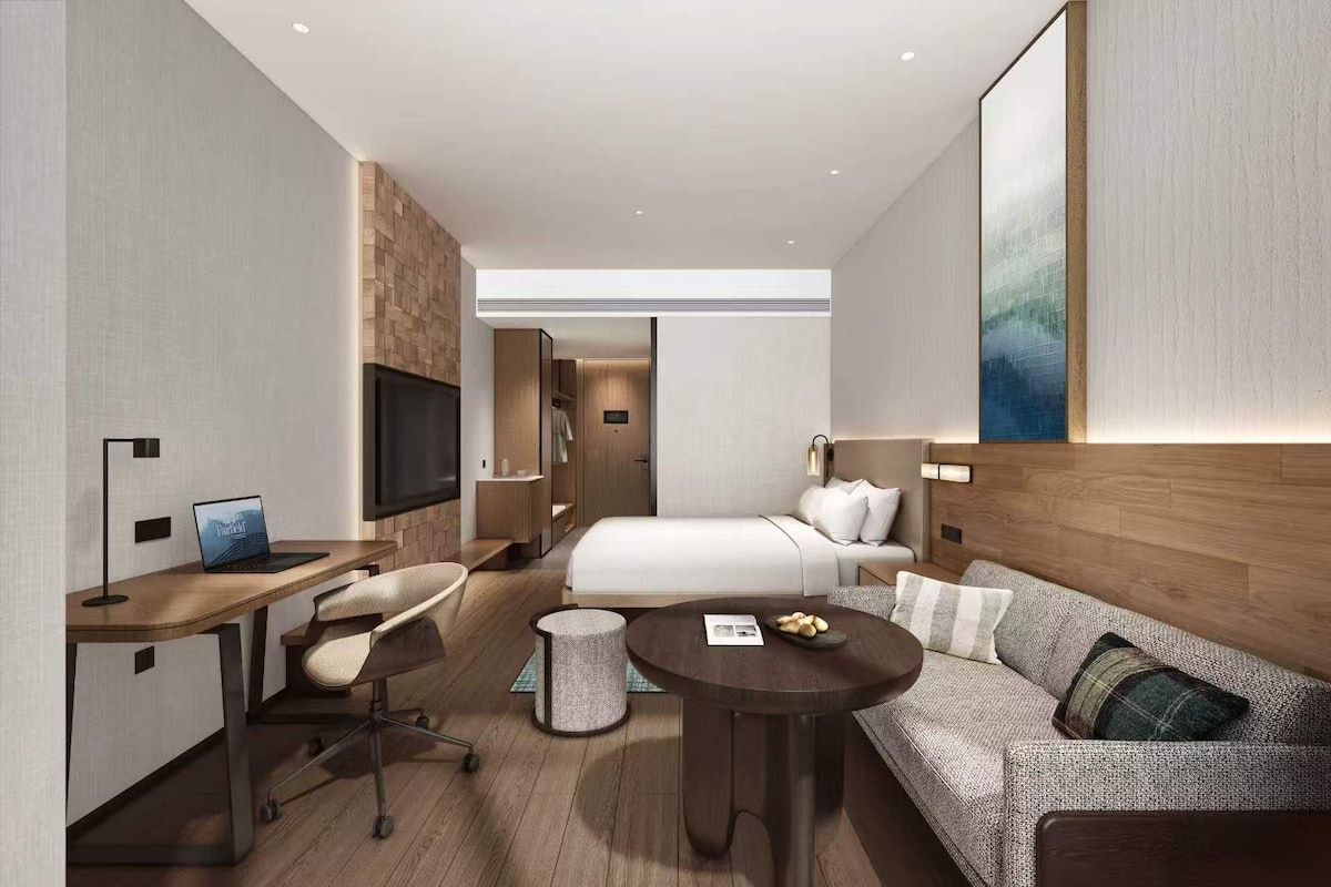 A modern hotel room with a bed, a sofa, a desk with a laptop, a TV, and a small round table with a plate of cookies.