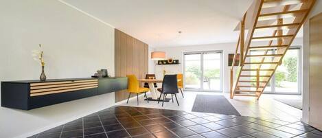 A modern interior with a black-tiled floor, a wooden staircase, a dining area with yellow chairs, and a black console table.