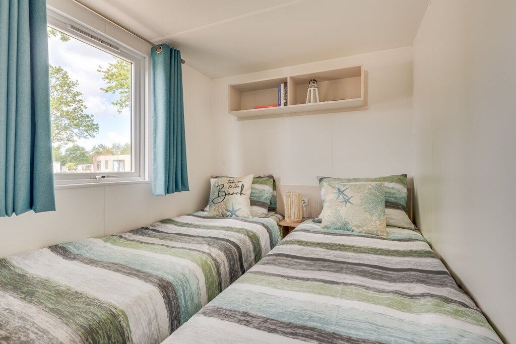 Un dormitorio con dos camas, una ventana con cortinas, un estante con libros y una lámpara, y una repisa en la pared sobre las camas.