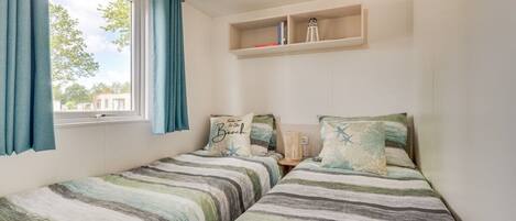 Un dormitorio con dos camas, una ventana con cortinas, un estante con libros y una lámpara, y una repisa en la pared sobre las camas.