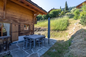 Eine Holzhütte mit einer Steinterrasse, Sitzgelegenheiten im Freien und Blick auf einen Hügelhang.