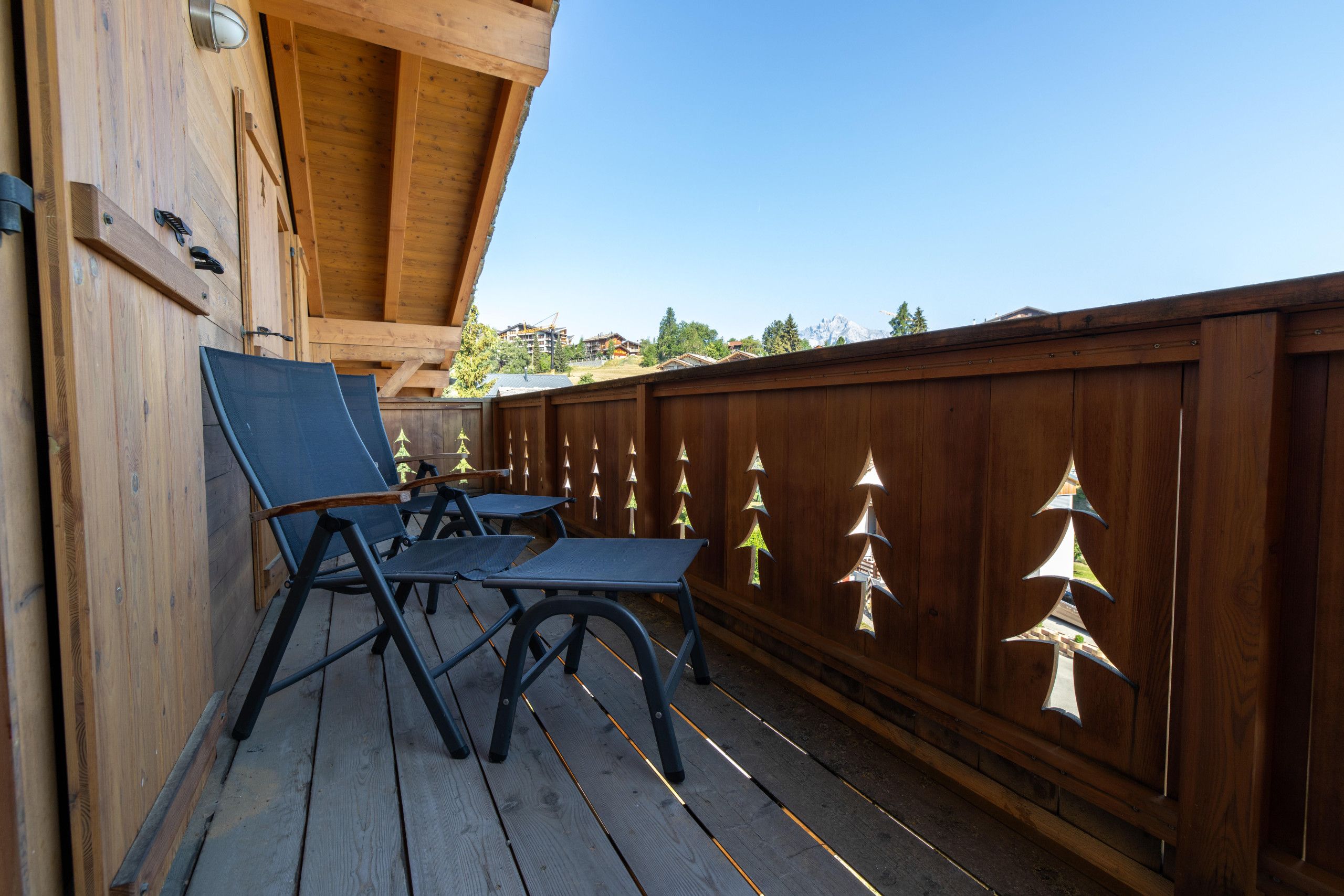 Une terrasse en bois avec deux chaises longues et une table, une clôture en bois ornée de motifs décoratifs, et un ciel dégagé.