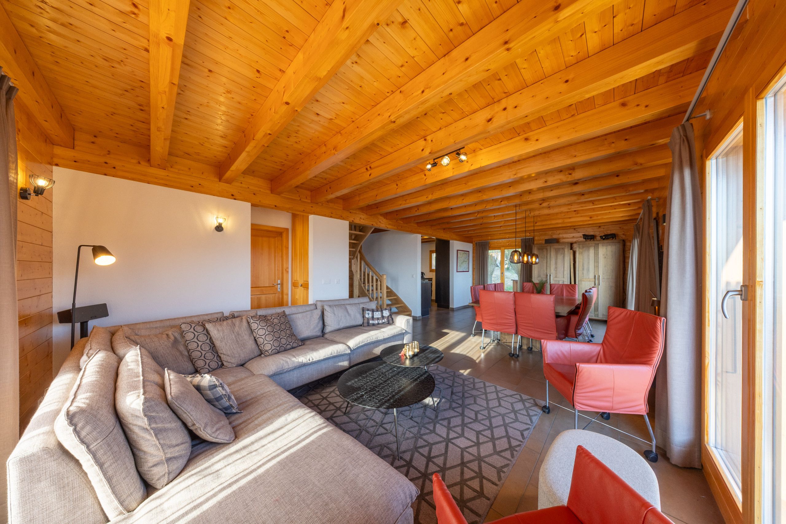 Ein geräumiger Wohnbereich mit Holzbalkendecke, einem Ecksofa, einem Couchtisch und einem Essbereich mit roten Stühlen.