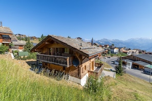 Eine Holzchalet mit einem Balkon, umgeben von Hügellandschaft und freiem Blick auf ferne Berge.