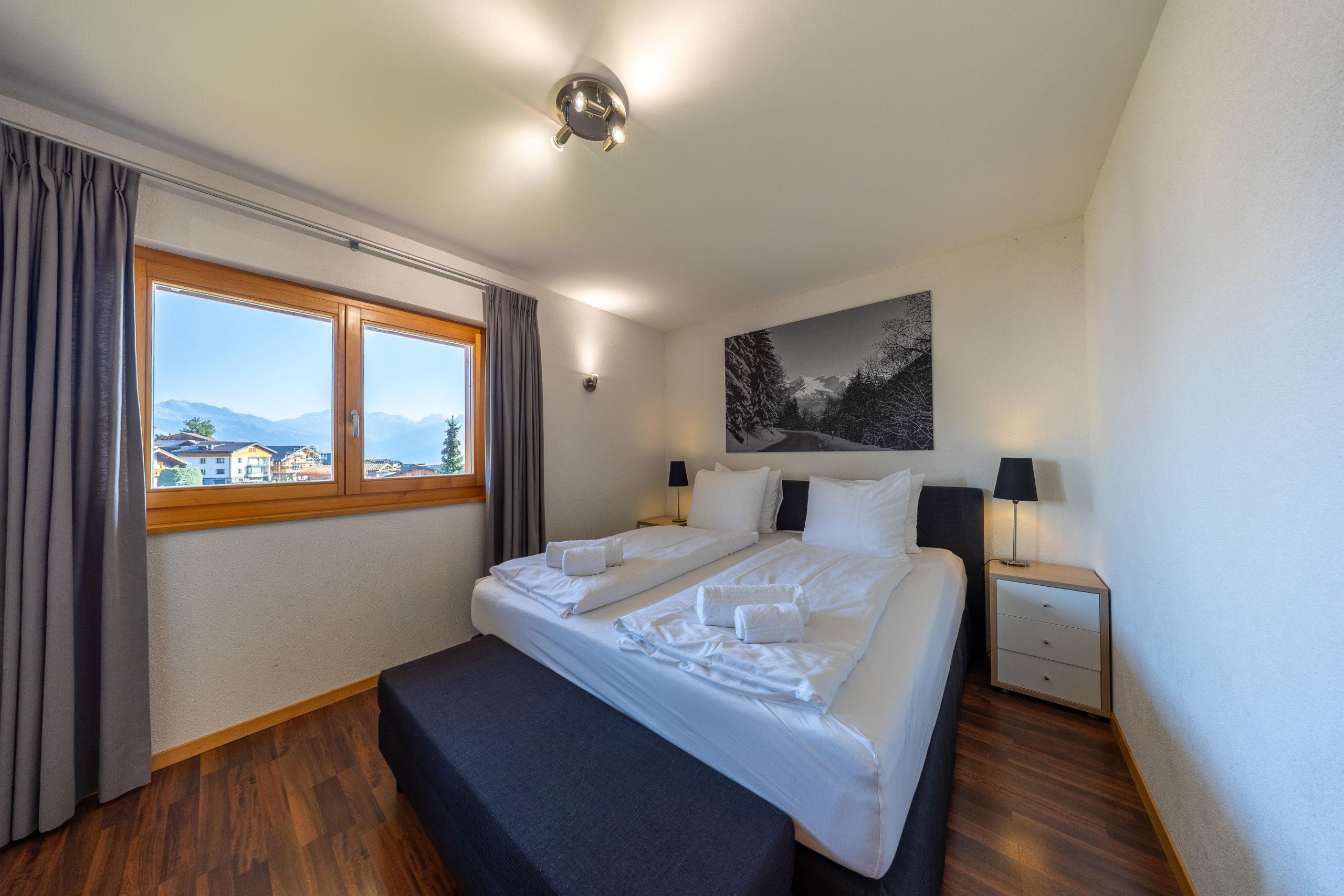 A hotel room with a bed, a bedside table, a lamp, a window with curtains, and a painting on the wall.