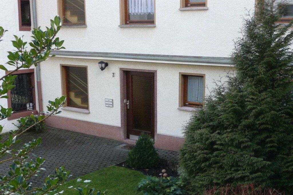 Un edificio de dos pisos con puerta principal, ventanas y un pequeño jardín con un árbol y arbustos.