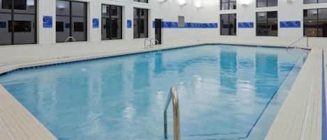 An indoor swimming pool with clear water, surrounded by a tiled deck and a white wall with windows.