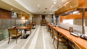 Uma sala de jantar de hotel com mesas e cadeiras de madeira, uma área de cozinha com eletrodomésticos de aço inoxidável e piso ladrilhado.