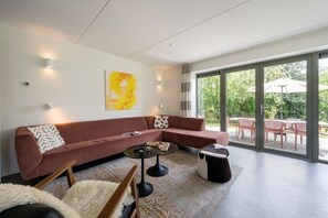 A modern living room with a sectional sofa, a coffee table, and a rug. There is a large window with a view of greenery outside.