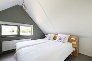 Ein Schlafzimmer mit schräg versiegelter Decke, einem großen Fenster, einem Bett mit weißer Bettwäsche und einem Holz-Kopfteil.