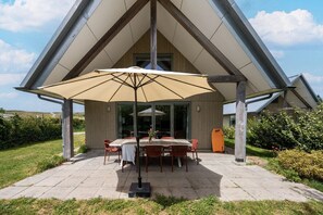 An outdoor dining area with a table and chairs under a large umbrella, surrounded by a wooden structure with a sloped roof.