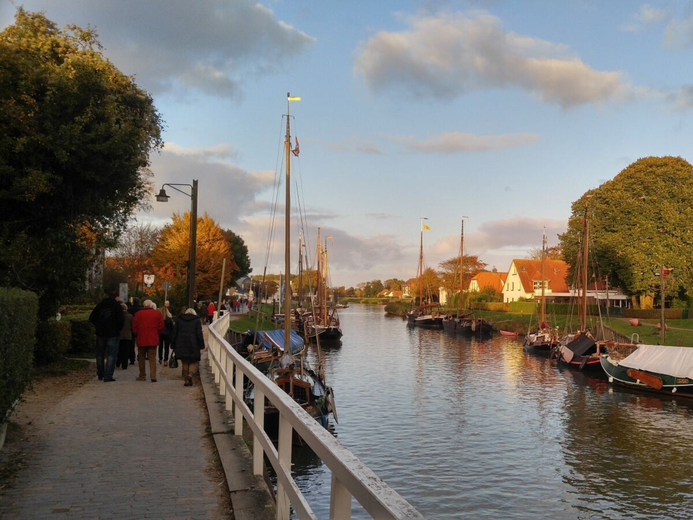 A riverside walkway with boats docked on the river, a white fence, and a building with a red roof in the background.