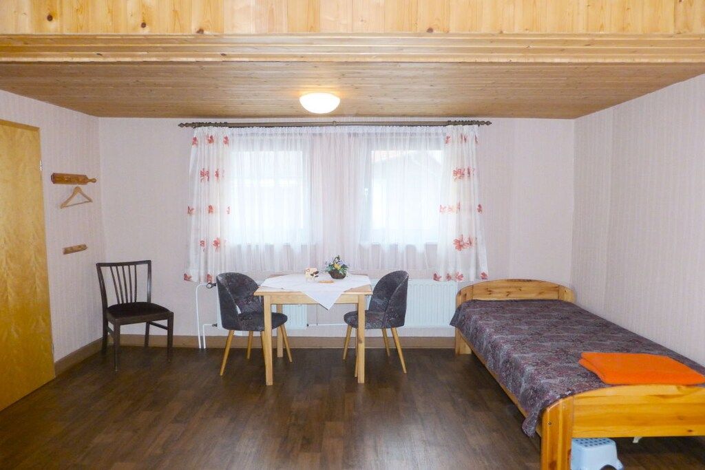 Een kamer met een houten plafond, een bed, een tafel met twee stoelen en een raam met gordijnen.