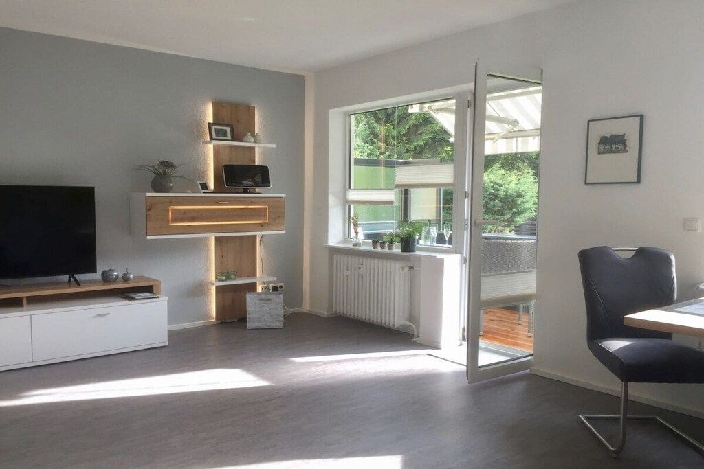 A modern living room with a flat-screen TV, wooden shelving, a radiator, and a large window with a view of greenery.