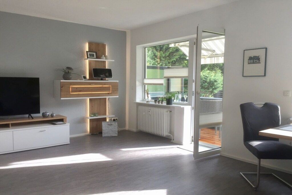 A modern living room with a flat-screen TV, wooden shelving, a radiator, and a large window with a view of greenery.