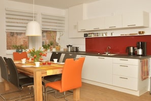 A modern kitchen with white cabinets, a red backsplash, and a dining area with a wooden table and orange chairs. - Holiday home with 2 bedrooms (Frankenfeld)