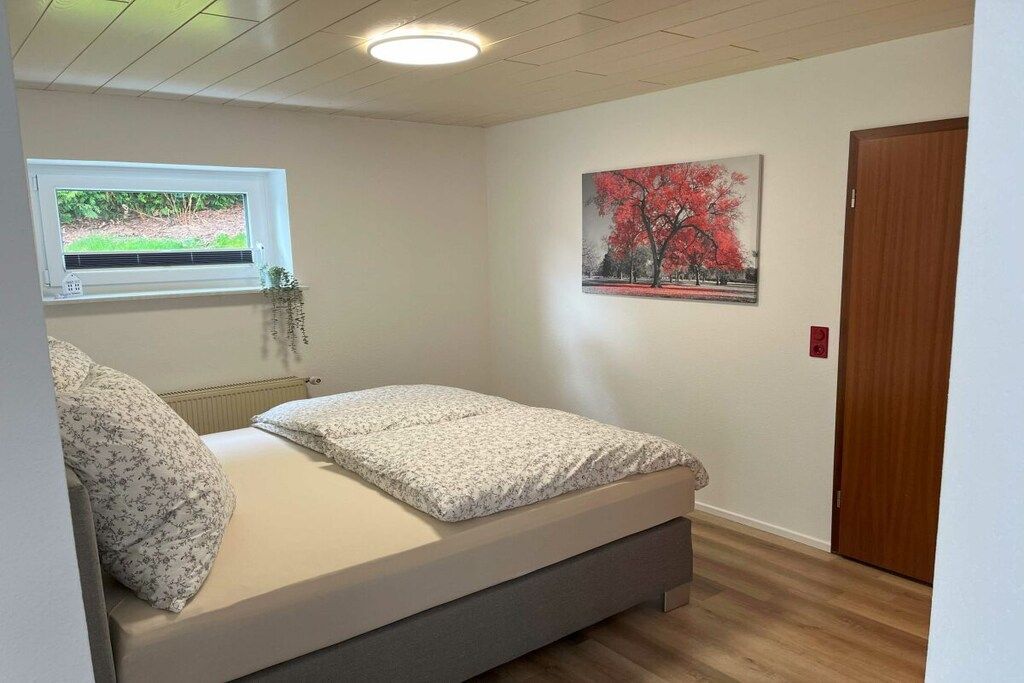 A bedroom with a bed, a window with a view, a painting on the wall, and a door.