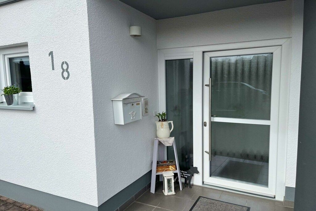 Une entrée d’habitation avec une porte blanche, une boîte aux lettres, une petite lanterne blanche et un pot de fleurs accroché au mur.