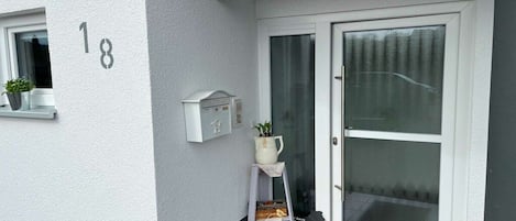 Une entrée d’habitation avec une porte blanche, une boîte aux lettres, une petite lanterne blanche et un pot de fleurs accroché au mur.