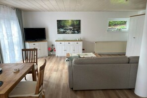 A living room with a television, sofa, dining table, and a painting of a forest stream. - Holiday apartment in Hünstetten with Wi-Fi (Hünstetten)