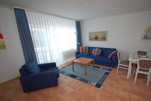 A living room with a sofa, a coffee table, a dining table with chairs, and a large window with curtains. - Schmidt - Mühlenstr. (Prerow)
