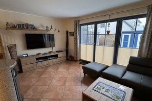 Ein Wohnzimmer mit einem Flachbildfernseher, einem Sofa, einem Couchtisch und einem großen Fenster mit Jalousien.
