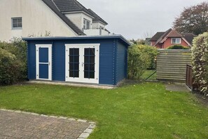 Eine blaue Gartenhütte mit weißen Fensterrahmen und einer Tür, gelegen in einem grünen Hinterhof mit einem Ziegelweg, der zu ihr führt.