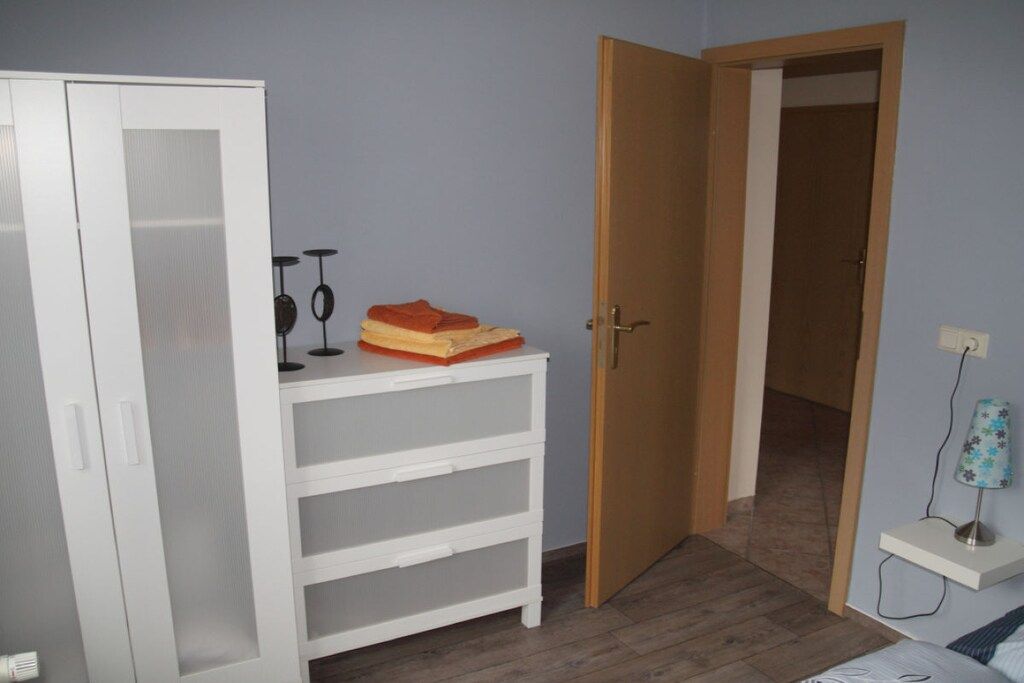 Una habitación con un armario blanco, una mesita pequeña con una lámpara y una puerta de madera que da a otra estancia.