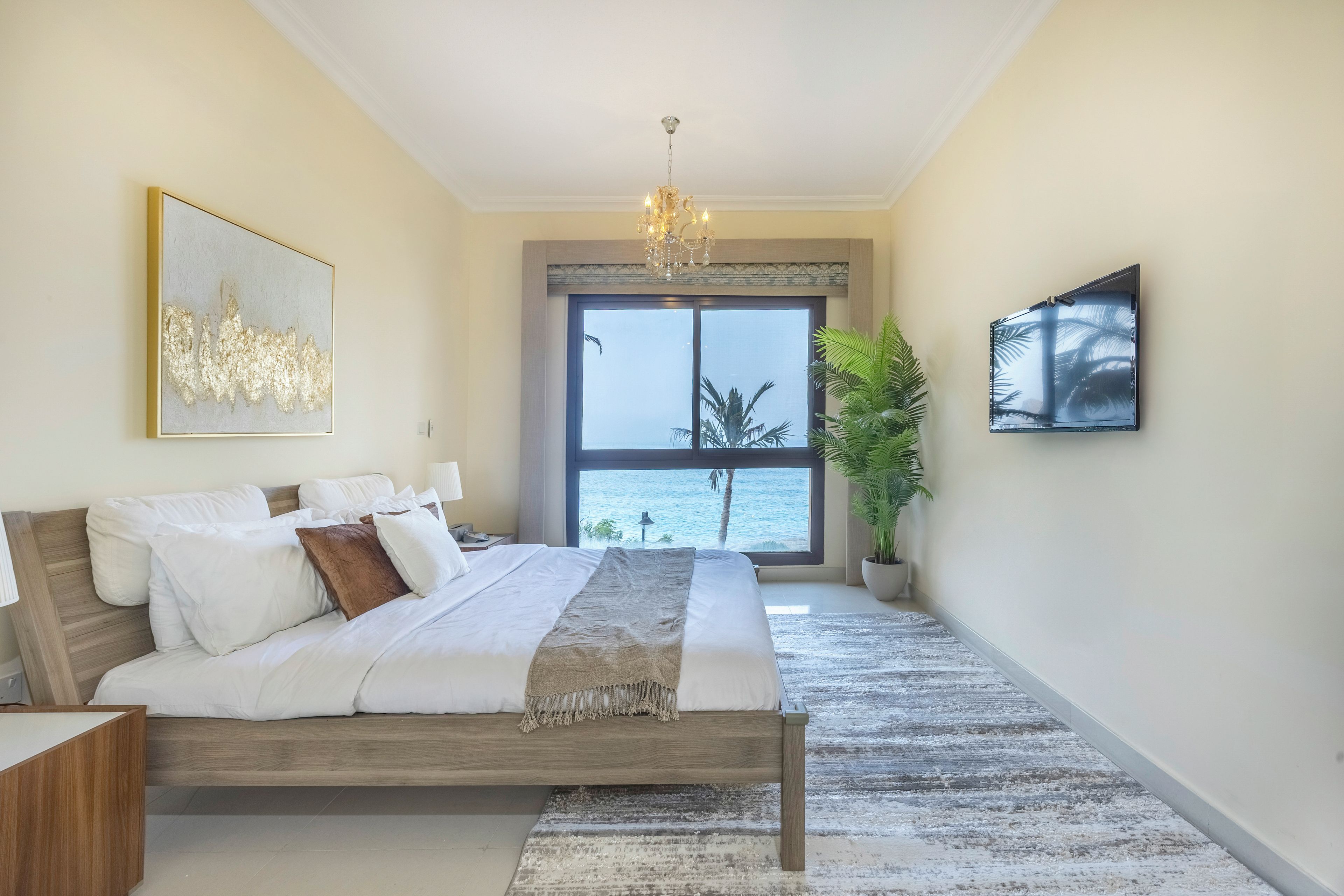 A bedroom with a large bed, a chandelier, a view of the sea, and a potted plant.