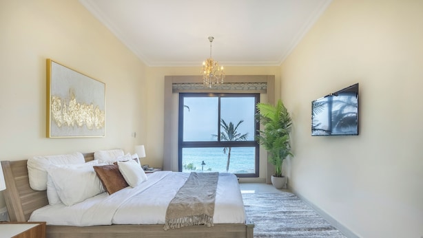 Ein Schlafzimmer mit einem großen Bett, einer Kristallleuchtre, Blick aufs Meer und einer Topfpflanze.