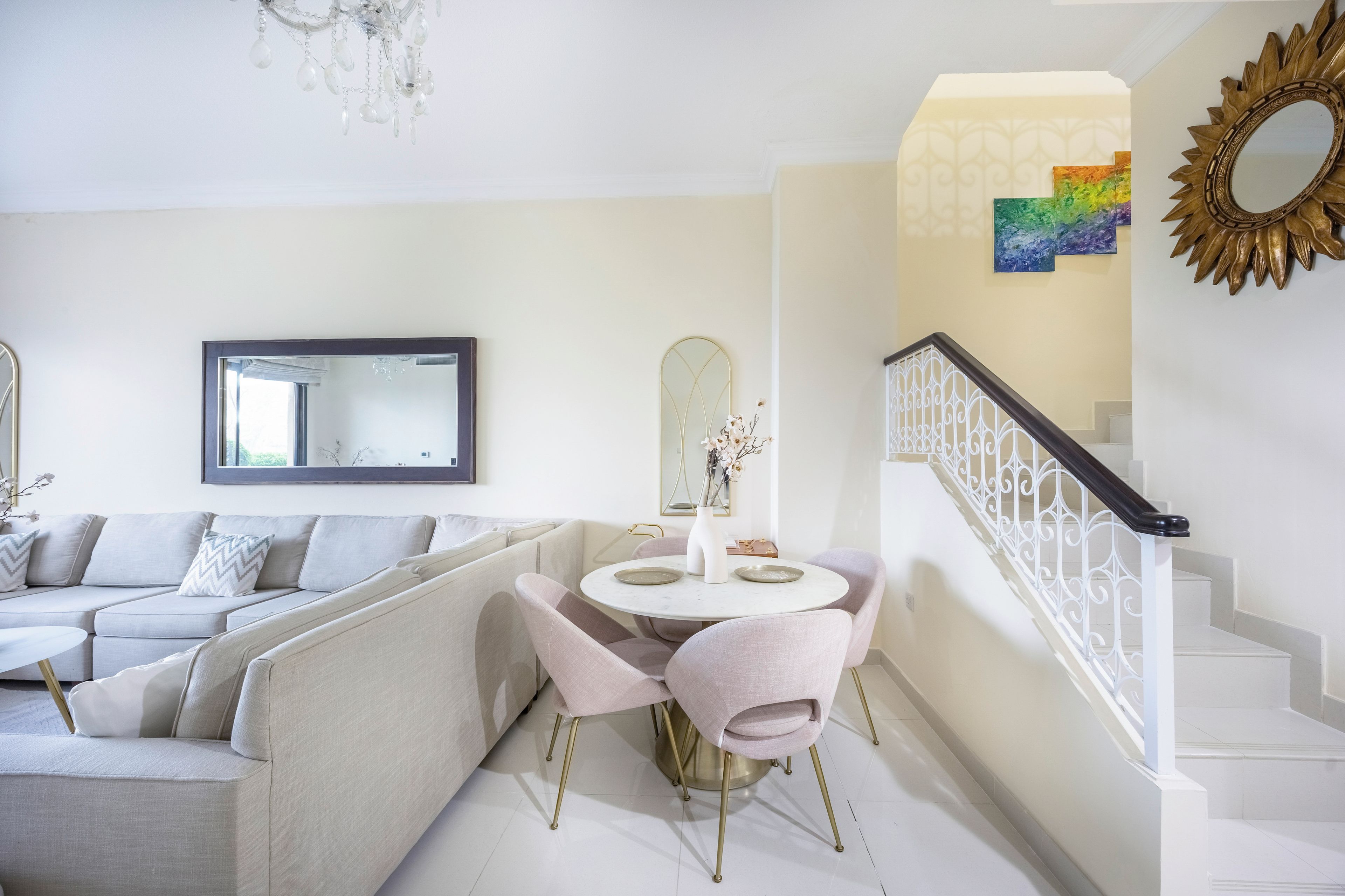 A modern living room with a sofa, a dining table with chairs, a staircase with a railing, and a chandelier.