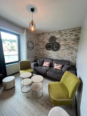 A modern living room with a grey sofa, green armchairs, and a coffee table. A decorative wall clock and a hanging light fixture are visible. - Harbor Master's Office—stunning harbor view—downtown (Port-en-Bessin-Huppain)