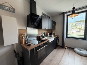 A modern kitchen with a built-in oven, microwave, and a large window offering a view of a residential area. - Harbor Master's Office—Breathtaking Harbor View—Downtown (Port-en-Bessin-Huppain)