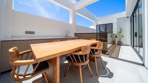 Un área de comedor exterior con mesa y sillas de madera, una cocina con fregadero y plantas en macetas.