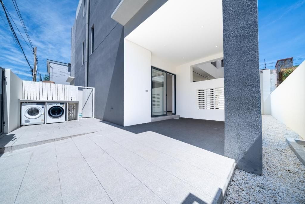 Un espace extérieur moderne doté d’un sol en béton, d’un lave-linge et d’un bâtiment à l’extérieur blanc et orné d’un pilier foncé.