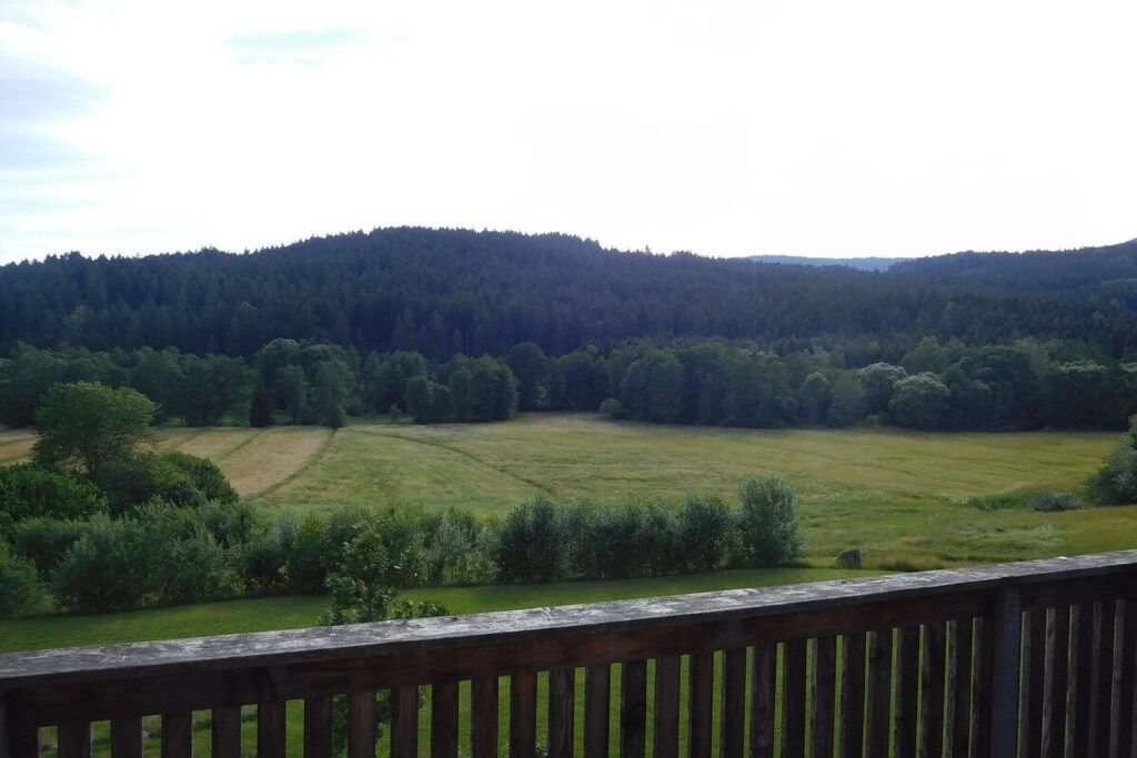 Vista desde una terraza de madera con vista a un campo cubierto de hierba y una colina boscosa al fondo.