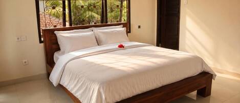 A neatly made bed with a white duvet and pillows, a wooden headboard, and a window with a view of greenery.