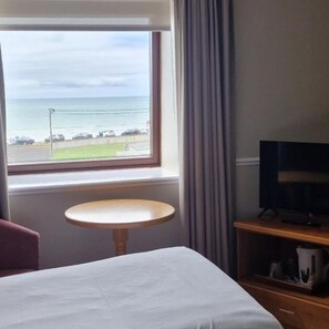 Quarto de hotel com cama, mesa redonda pequena, TV sobre suporte de madeira e janela com vista para o mar e uma estrada.