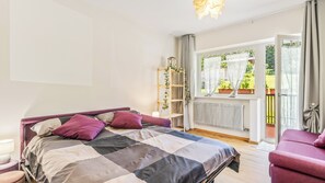Ein Schlafzimmer mit Bett, Nachttisch, Sessel, Heizkörper, Fenster mit Vorhängen und einem Balkon mit Pflanzen.